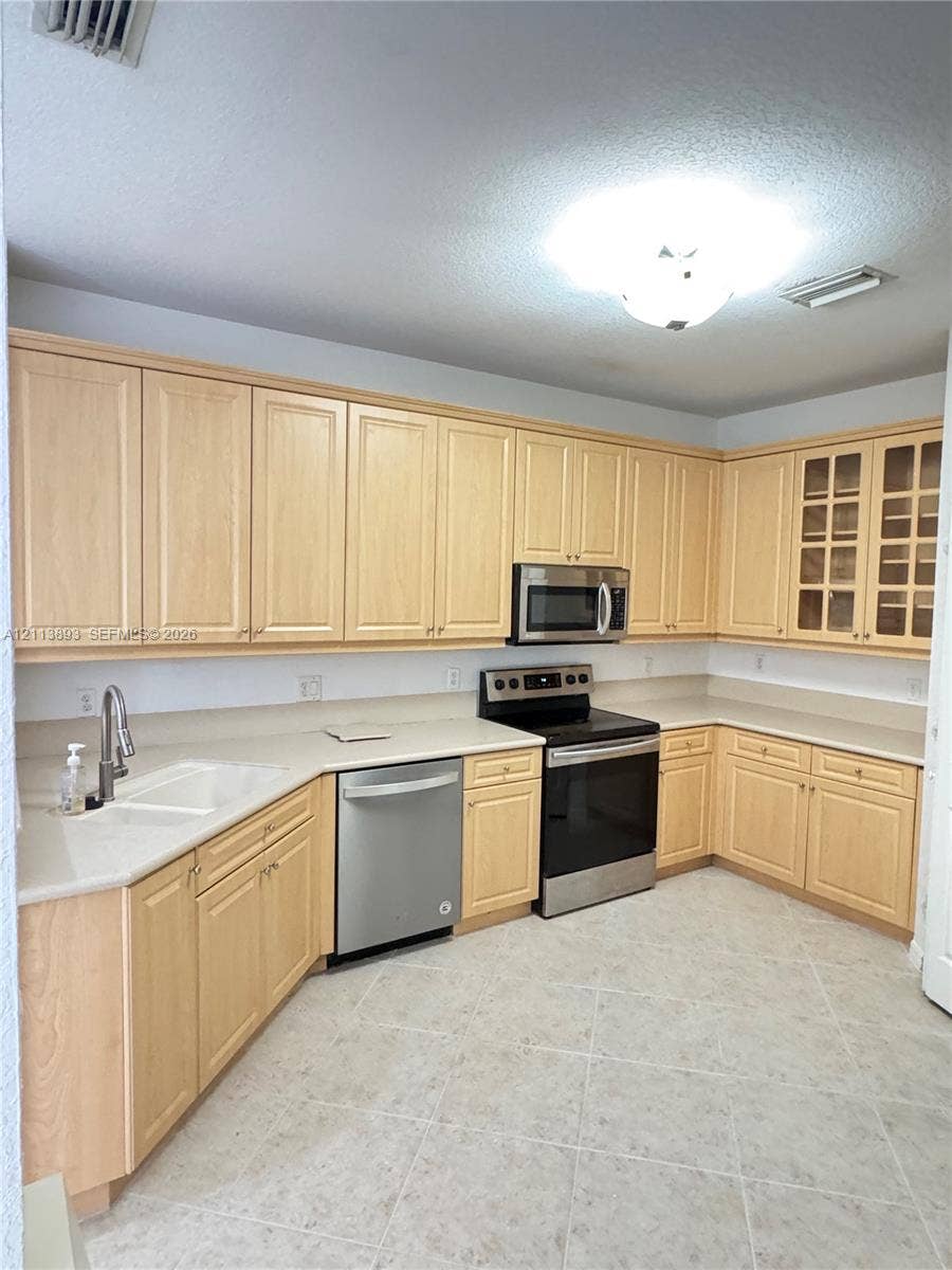 5737 NW 119th Drive, Coral Springs, FL 33076 Light-toned cabinetry with integrated glass-paneled display uppers, expansive solid-surface countertops, tile flooring, and stainless steel appliances including a built-in microwave