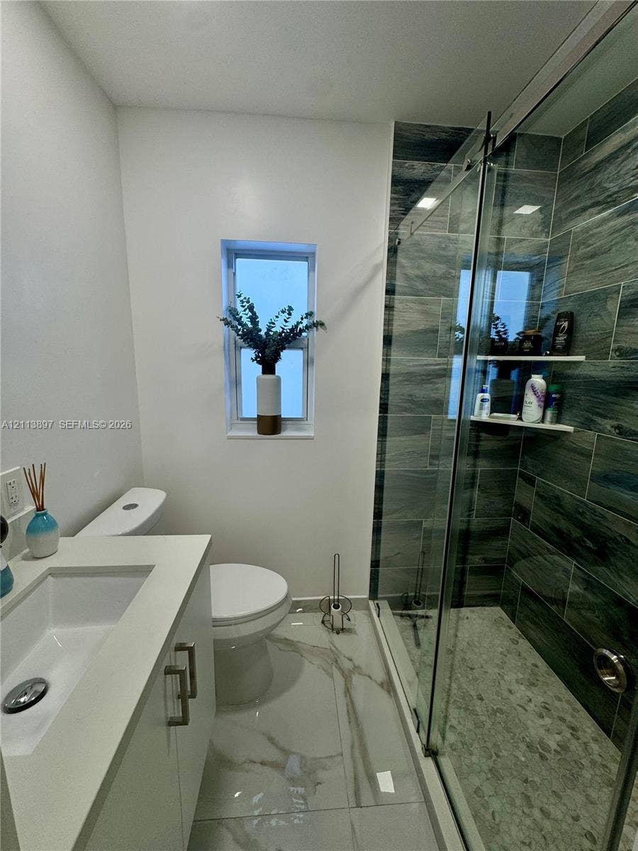520 E 58th Street, Hialeah, FL 33013 Bathroom featuring large-format stone-finish floor tiling, a vanity with a rectangular integrated sink, a walk-in shower with stone-textured wall tiles and a glass sliding door, and integrated shower shelving