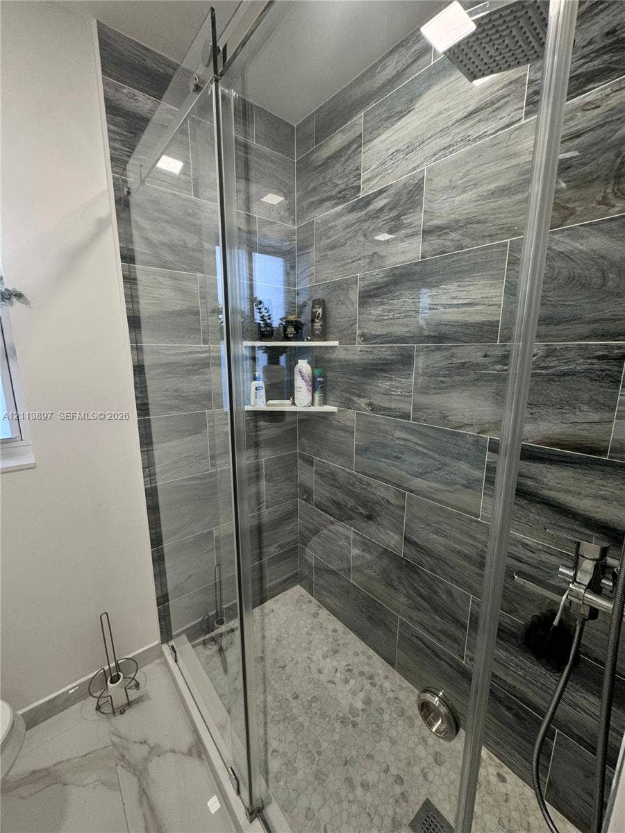 520 E 58th Street, Hialeah, FL 33013 Walk-in shower featuring large-format stone-pattern wall tile, pebble-texture floor tiling, a frameless glass sliding door, a built-in corner shelf, and a rainfall showerhead