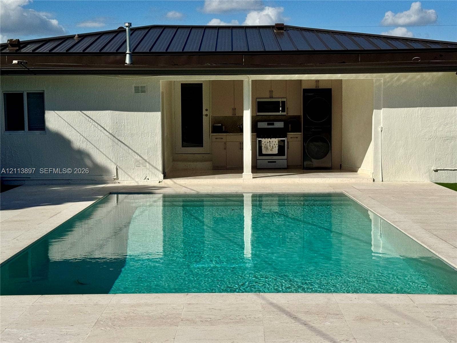 520 E 58th Street, Hialeah, FL 33013 Rectangular in-ground pool surrounded by light-toned stone decking, featuring a covered outdoor kitchen area with built-in cabinetry, stainless steel appliances, and a stacked washer and dryer set