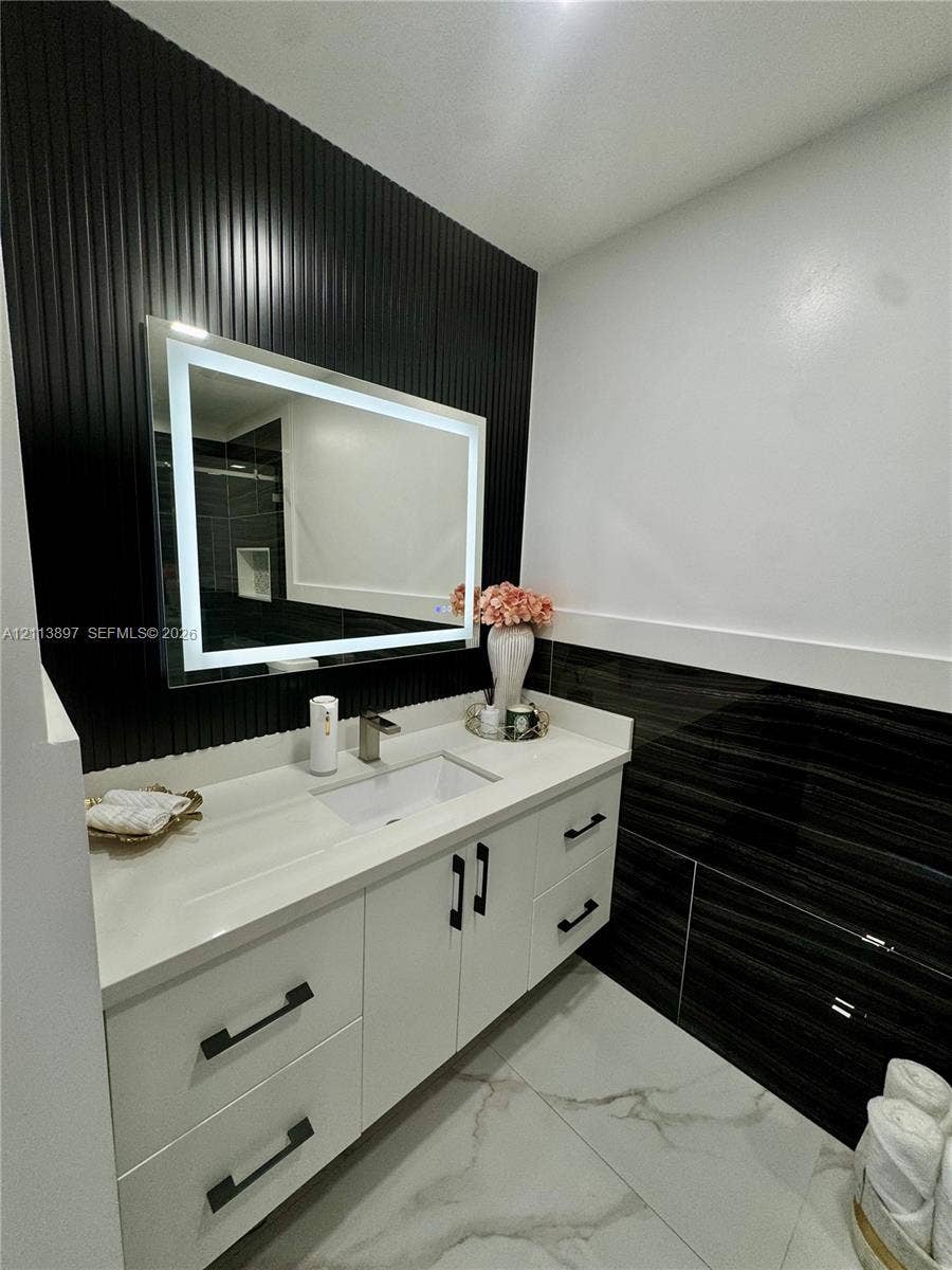 520 E 58th Street, Hialeah, FL 33013 White vanity with clean-lined cabinetry, quartz countertop, integrated rectangular sink, illuminated mirror, and vertical slat accent wall