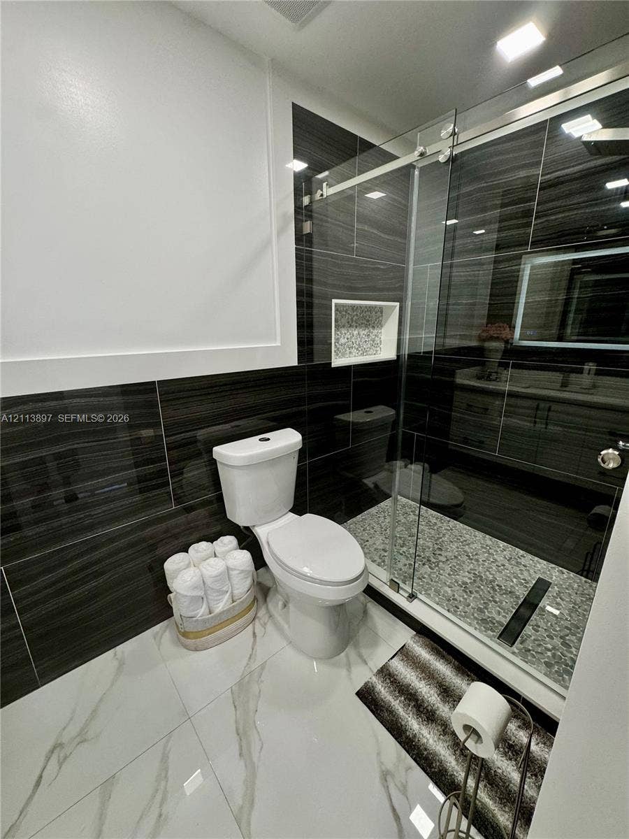 520 E 58th Street, Hialeah, FL 33013 Bathroom featuring large-format white tile flooring, dark horizontal-patterned wall tiling, a walk-in shower with a glass sliding door, a recessed shower niche, and a vanity cabinet with an integrated LED-lit mirror