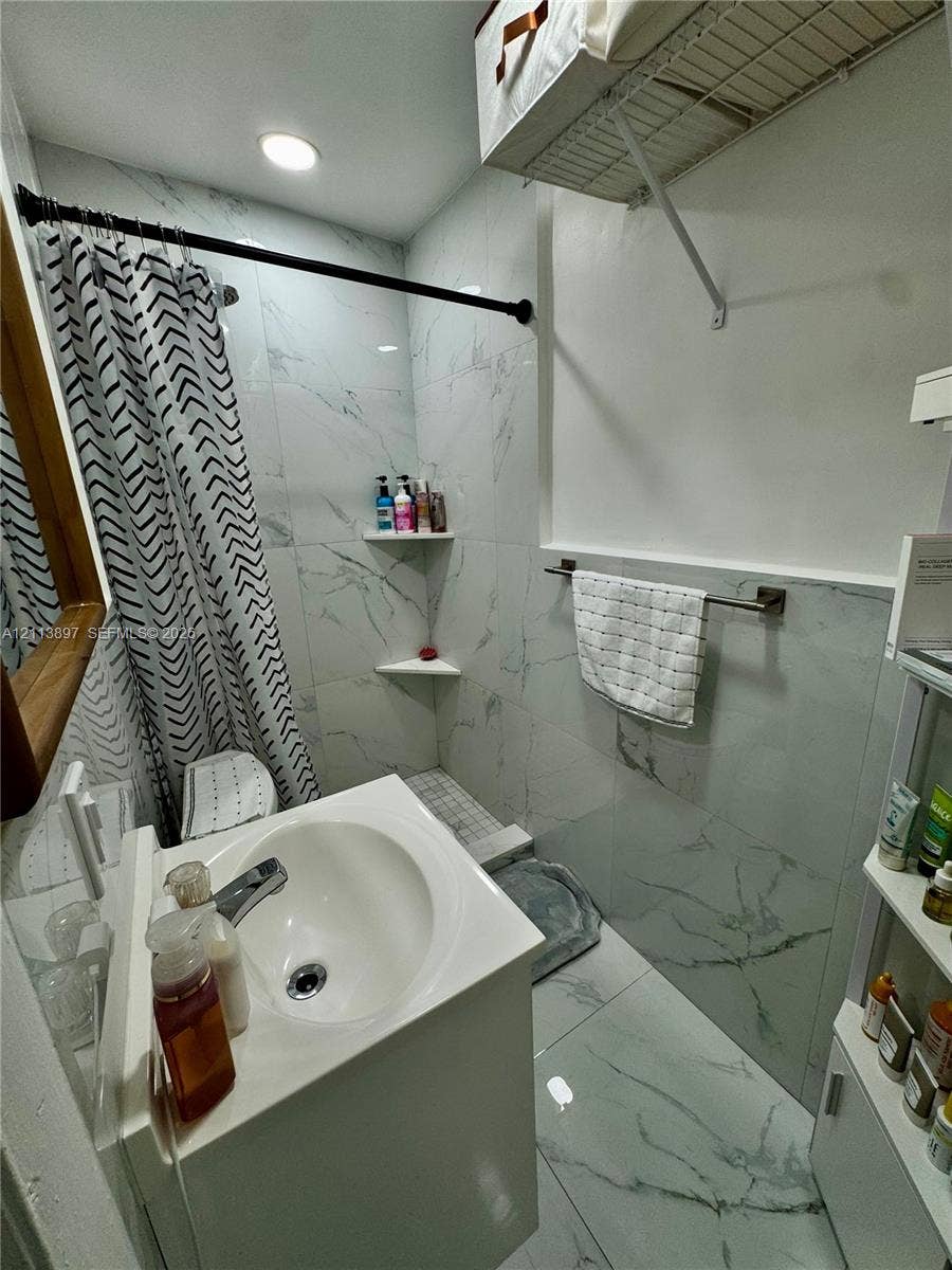 520 E 58th Street, Hialeah, FL 33013 Full-height tiled shower and floor surround with a white stone-pattern finish, recessed shelving, built-in wire storage rack, and wall-mounted metal towel bar