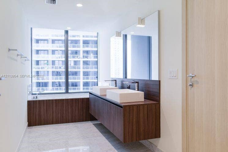 Floating wood-finish double vanity with dual square vessel sinks and chrome faucets, large frameless wall mirror, soaking tub with matching wood-finish surround, floor-to-ceiling windows, and light-toned stone-finish flooring