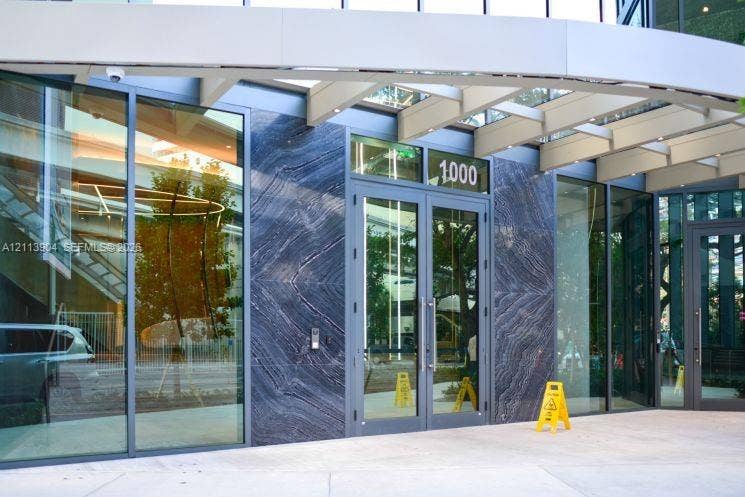 Building entrance featuring floor-to-ceiling glass paneling, dark stone facade cladding, a metal-framed canopy, and double-glass entry doors