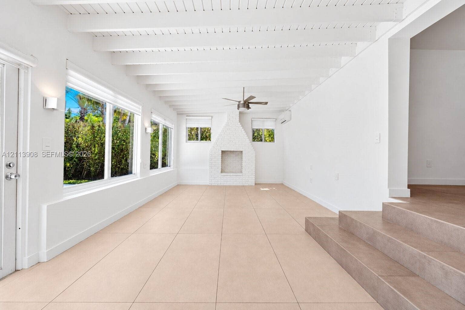 Vaulted ceiling with exposed white beams, large-format tile flooring, brick-surround fireplace, and multiple windows