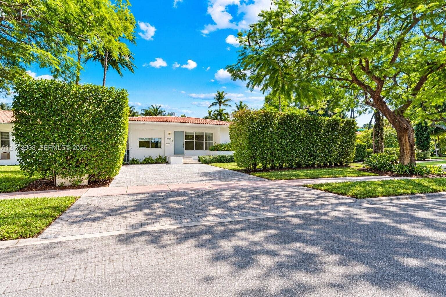 Single-story exterior featuring a clay tile roof, paver driveway, manicured privacy hedging, and mature canopy trees