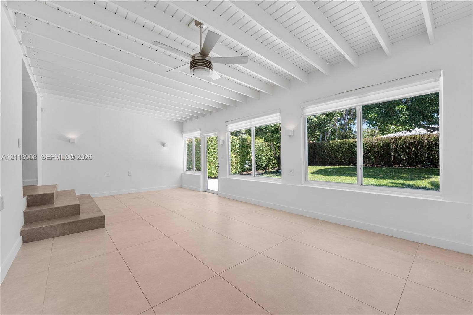 Open-concept space featuring expansive floor-to-ceiling windows, high exposed-beam ceilings, large-format tile flooring, and a central ceiling fan