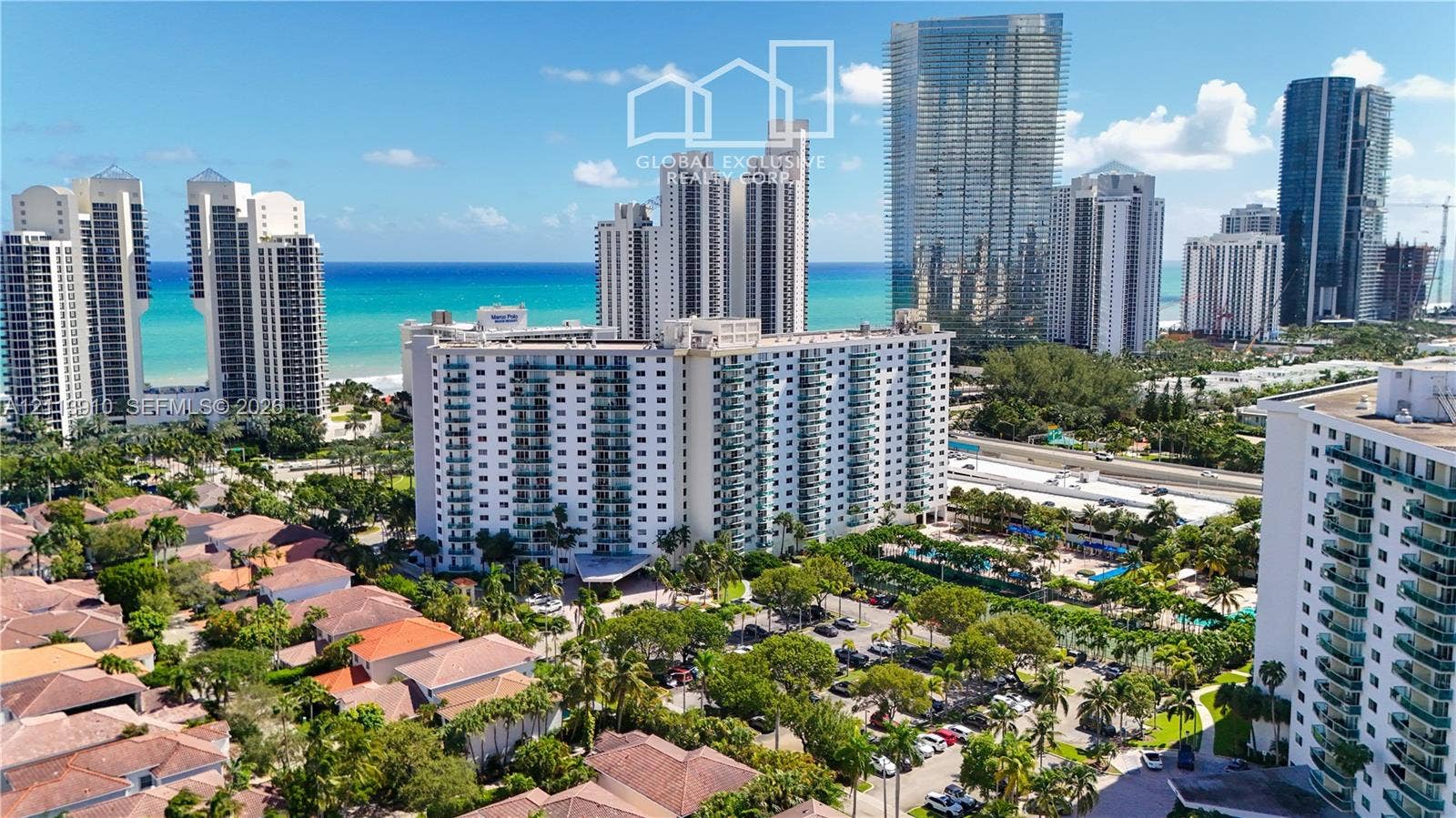 19380 Collins Avenue, #218, Sunny Isles Beach, FL 33160 Aerial perspective featuring a multi-story residential building with private balconies, surrounded by mature palm trees, paved parking areas, and proximity to the coastline
