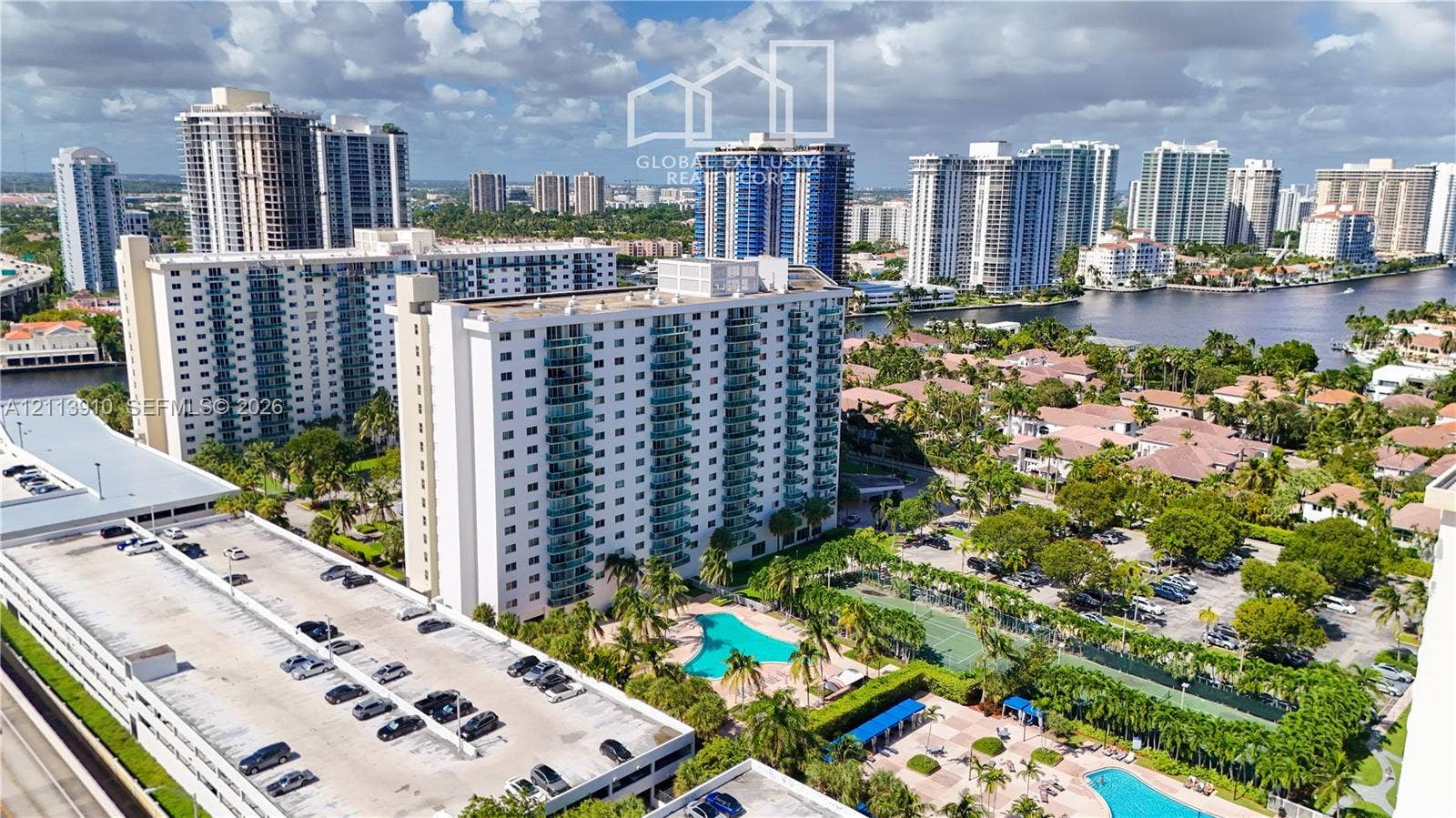 19380 Collins Avenue, #218, Sunny Isles Beach, FL 33160 Aerial perspective of a multi-story residential complex featuring an outdoor swimming pool, tennis courts, paved parking structures, and mature landscaping