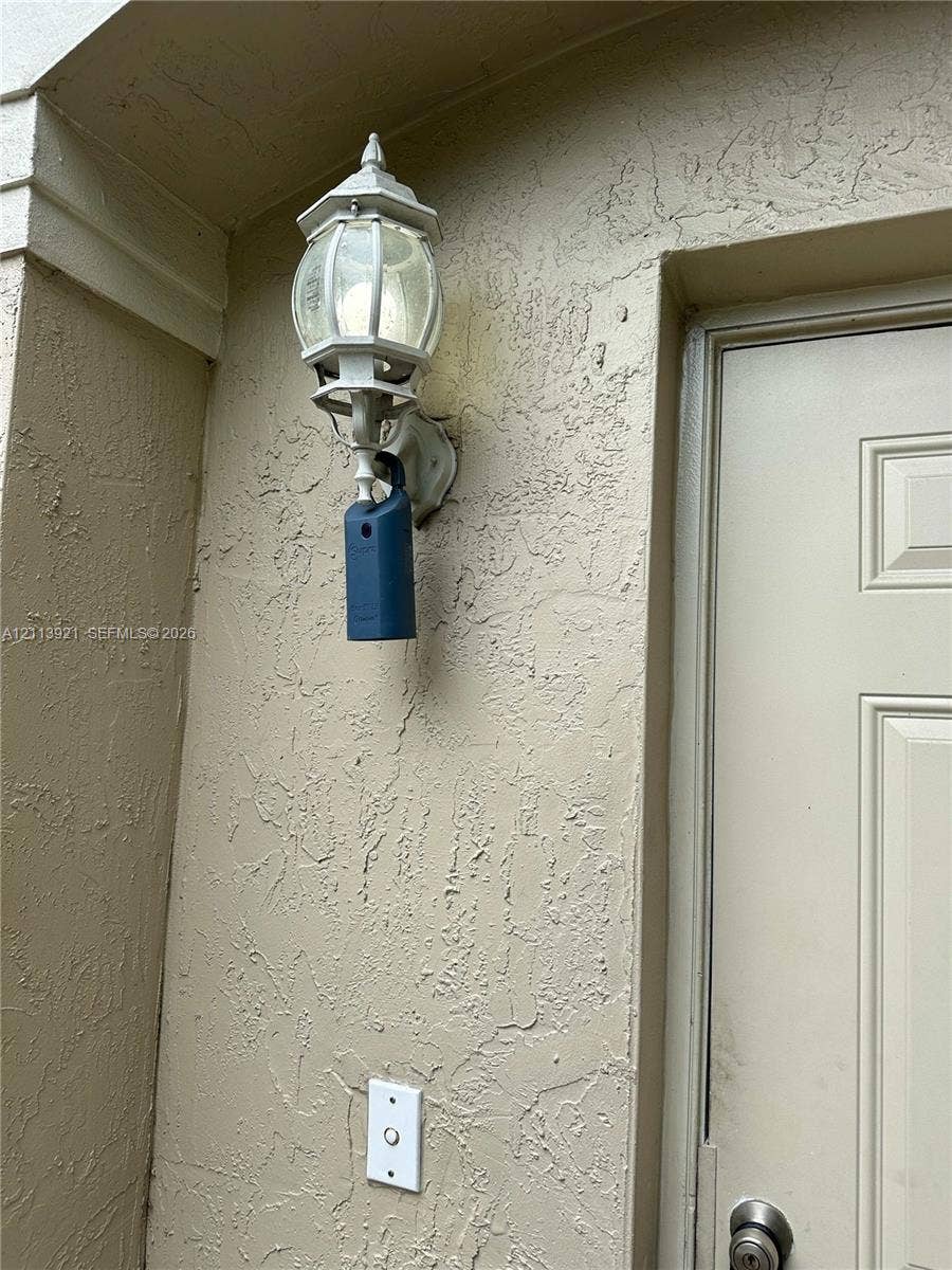824 Summit Lake Drive, West Palm Beach, West Palm Beach, FL 33406 Exterior wall featuring a textured stucco finish, traditional carriage-style wall-mounted light fixture, and a solid-panel door with a metallic hardware finish