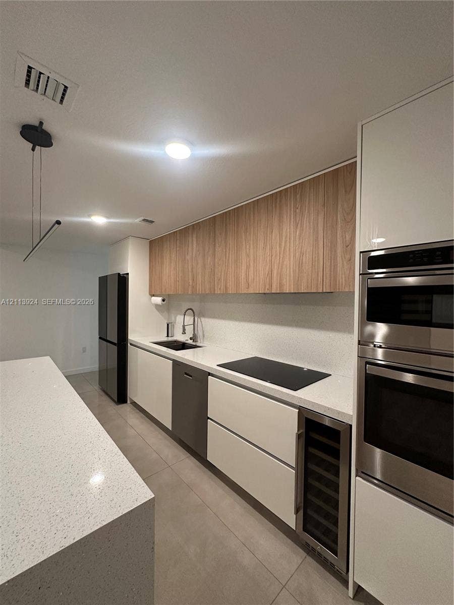 14569 SW 261st Terrace, #14569, Homestead, Homestead, FL 33032 Contemporary kitchen featuring a stone-surface waterfall island, flat-panel cabinetry with wood-finish upper units, integrated stainless steel appliances, built-in beverage refrigerator, and solid-surface countertops