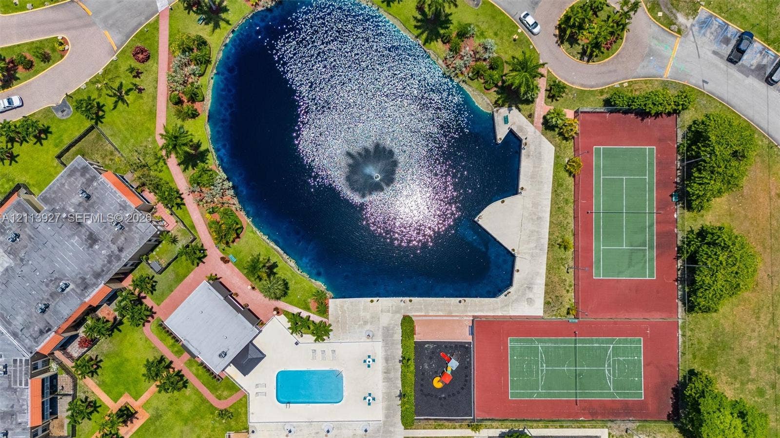 4900 NW 79th Avenue, #202, Doral, Doral, FL 33166 Aerial view featuring a central lake with a fountain, swimming pool, two hard-court surfaces, and paved walkways throughout the grounds