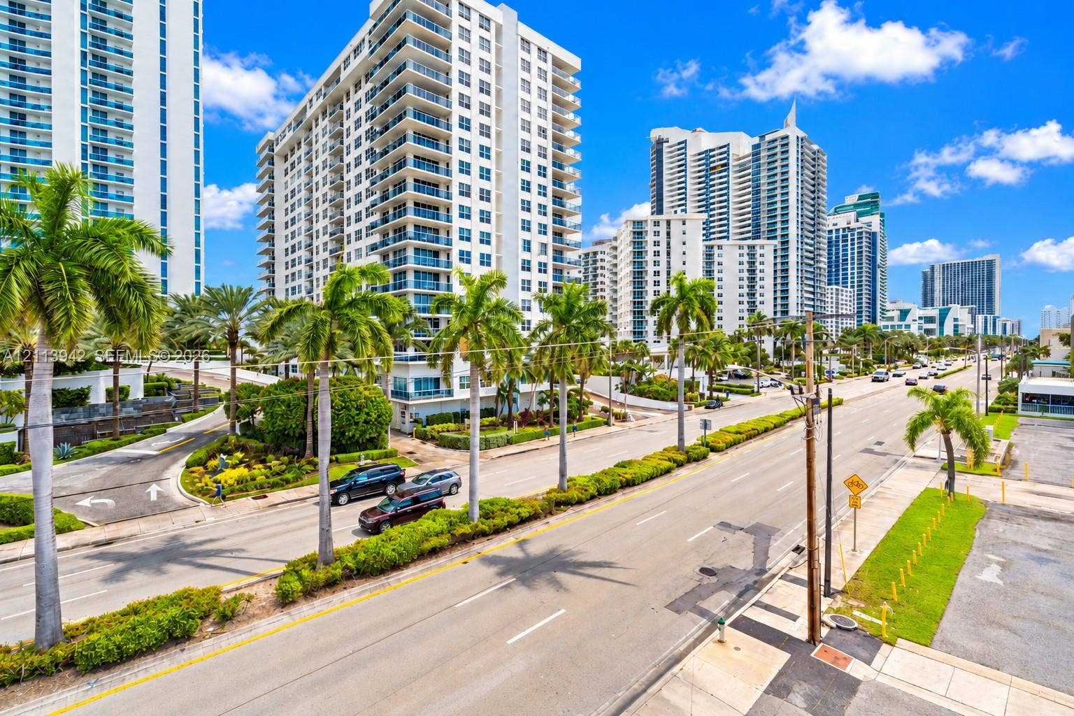 2710 S Ocean Drive, #401, Hollywood, FL 33019 High-rise residential exterior featuring glass balcony railings, white concrete facade, mature palm tree landscaping, and paved multi-lane street access