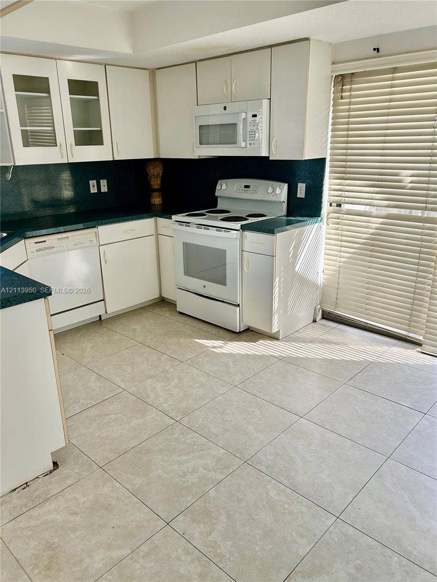 Kitchen featuring white cabinetry with glass-front uppers, solid surface countertops, square tile flooring, and integrated white appliances