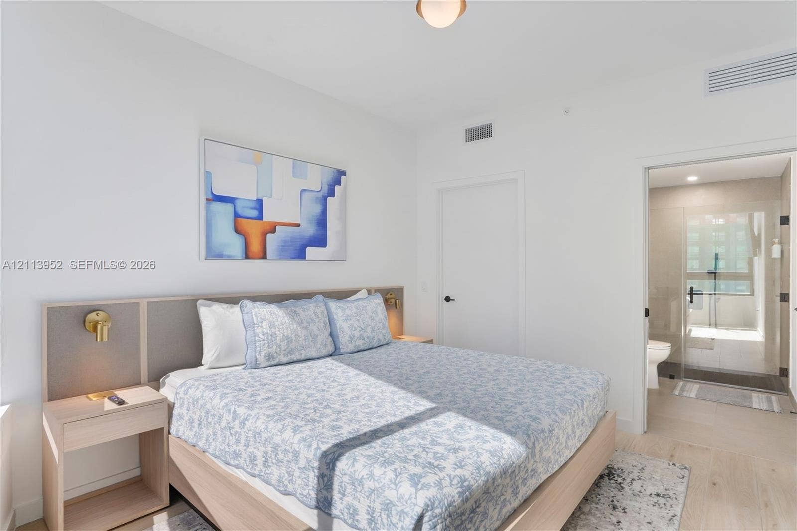 Neutral-toned bedroom featuring a built-in wood-finish headboard with integrated bedside tables, brass wall-mounted light fixtures, light-toned flooring, and a glass-enclosed shower stall