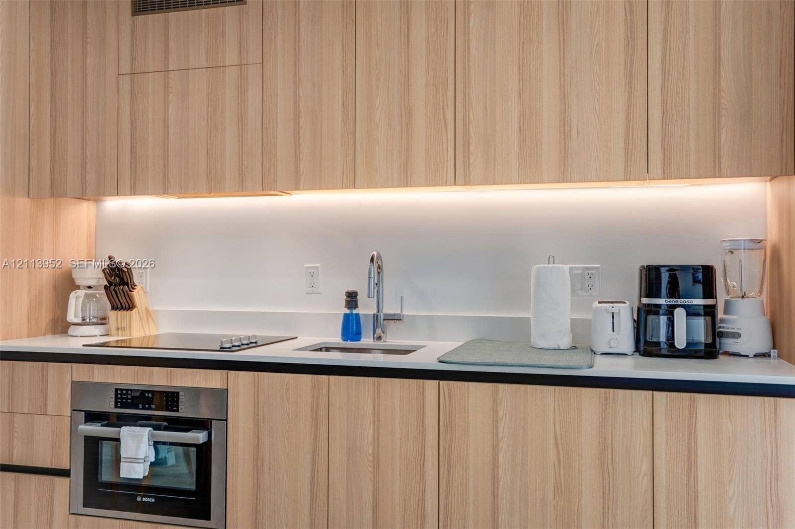 Kitchen featuring light wood-finish cabinetry, solid surface countertops, a flat-surface cooktop, an integrated stainless steel wall oven, and under-cabinet task lighting