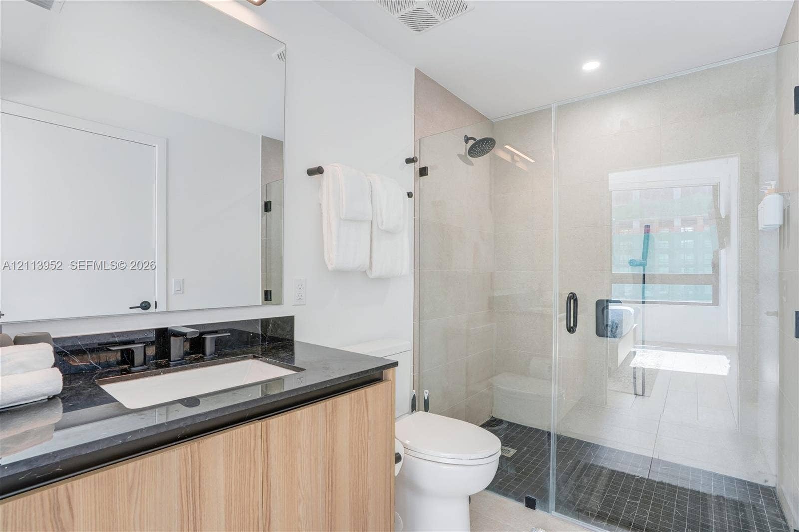 Bathroom featuring a vanity with light wood-finish cabinetry, dark stone countertop, integrated sink, frameless glass-enclosed shower with a rainfall showerhead, and a wall-mounted mirror