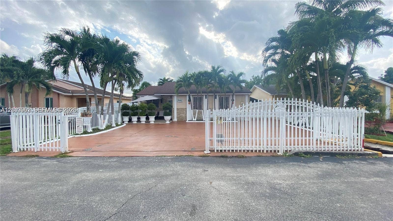 19764 NW 54 Avenue, Miami Gardens, FL 33055 Exterior facade featuring a paved driveway, a white perimeter fence with gated access, and mature palm trees