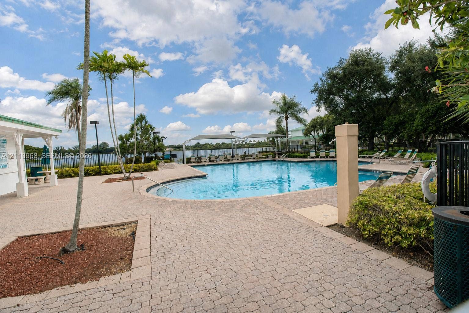 In-ground pool with paved surround, covered patio structure, and waterfront backdrop