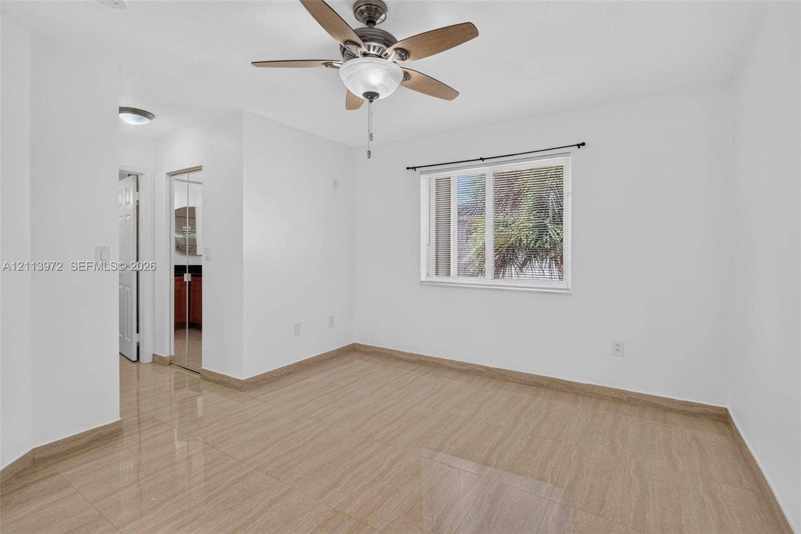 Neutral interior room featuring wood-finish tile flooring, a multi-blade ceiling fan with integrated lighting, a sliding window with black curtain hardware, and mirrored closet doors