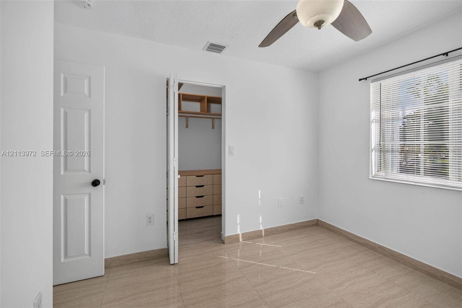 Polished tile flooring, neutral wall finishes, ceiling fan with integrated lighting, window with horizontal blinds, and walk-in closet featuring built-in shelving and drawer storage