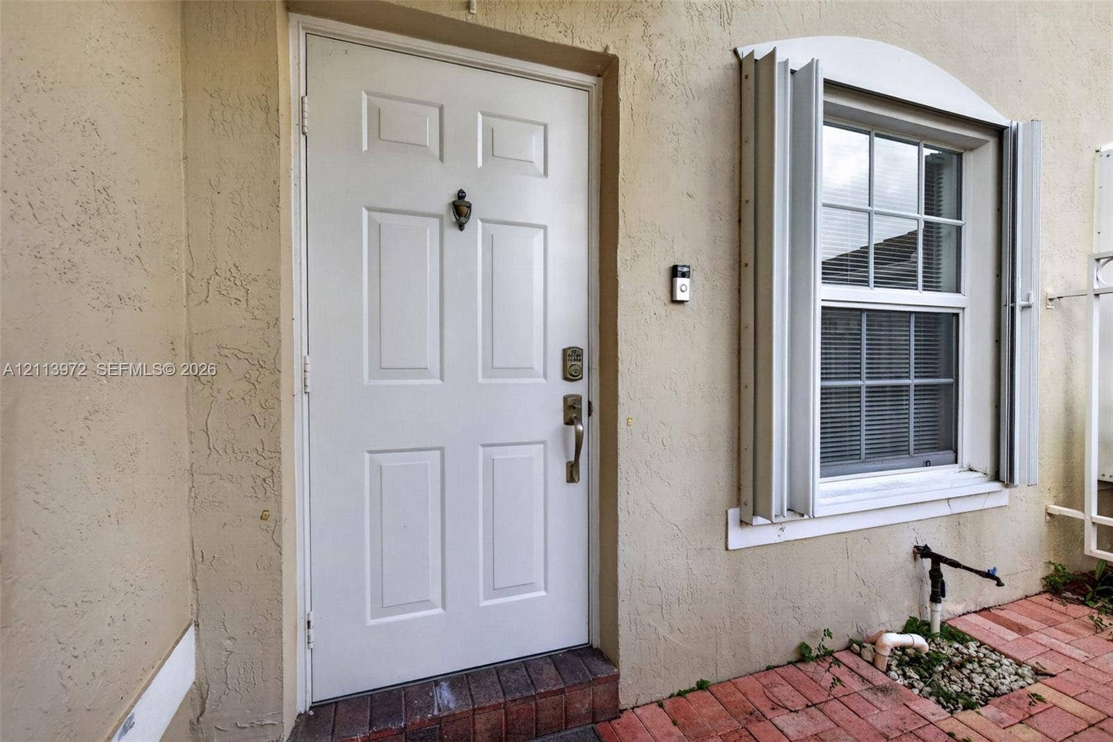 Exterior entryway featuring a six-panel white door, arched window with metal shutters, textured stucco siding, and a red brick patio