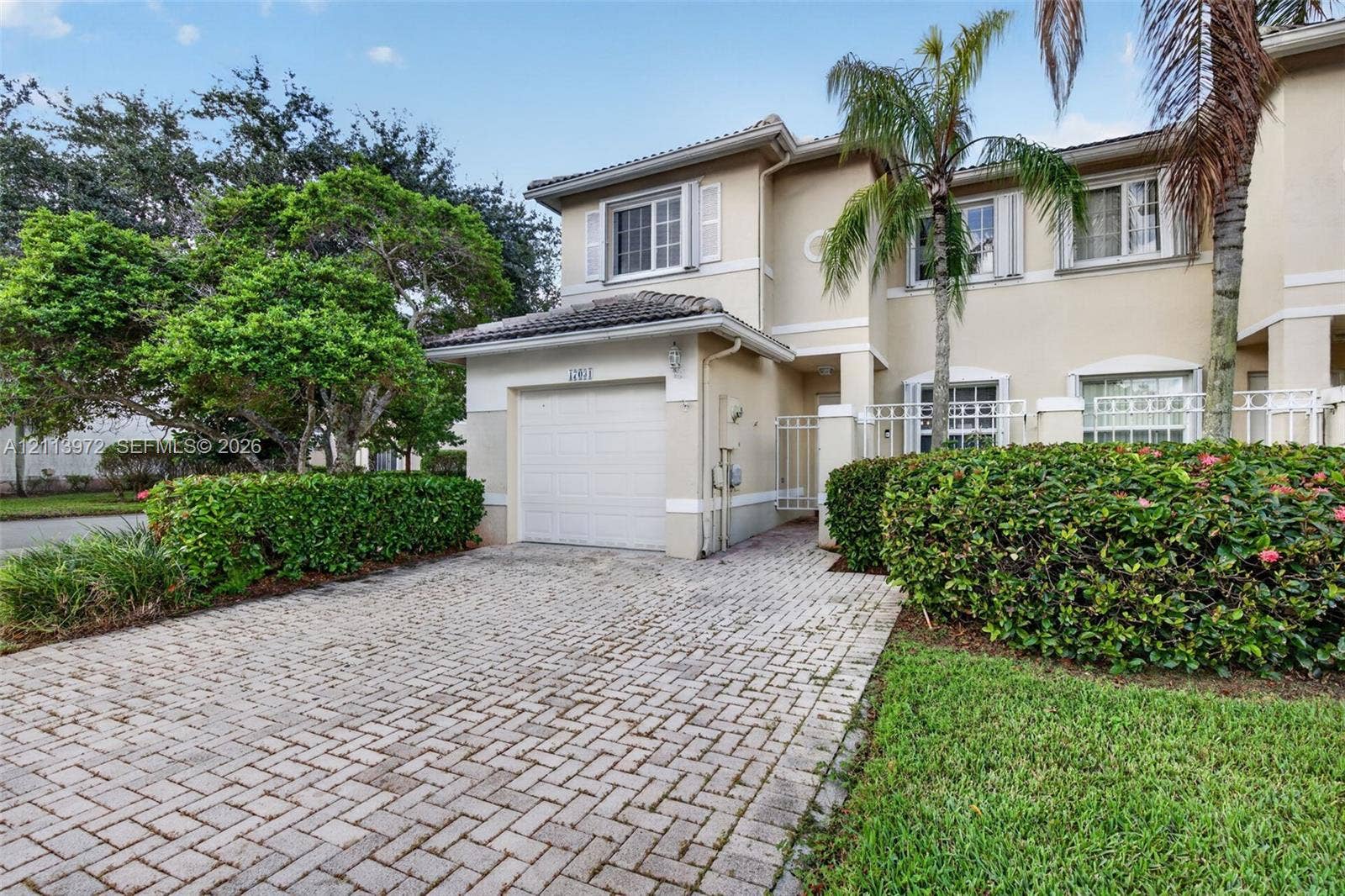 Two-story stucco exterior featuring a paver driveway, single-car garage, manicured landscaping, and decorative shutters