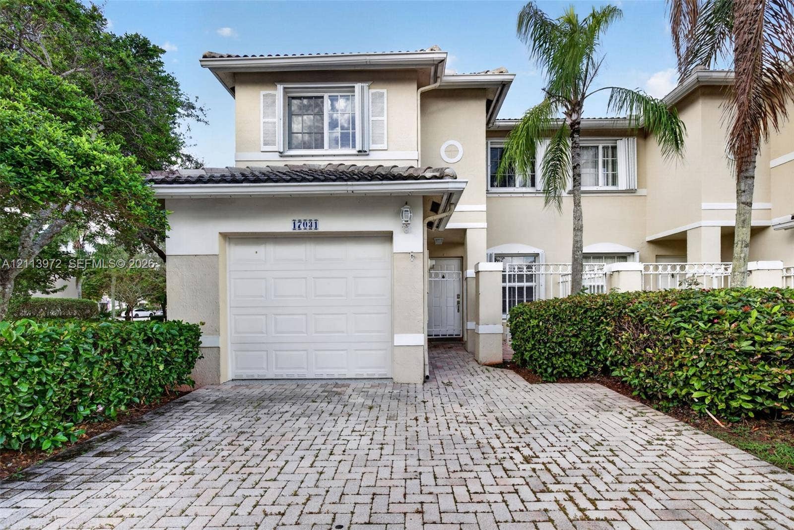 Two-story stucco exterior featuring a tiled roof, attached single-car garage, brick-paver driveway, and manicured perimeter landscaping