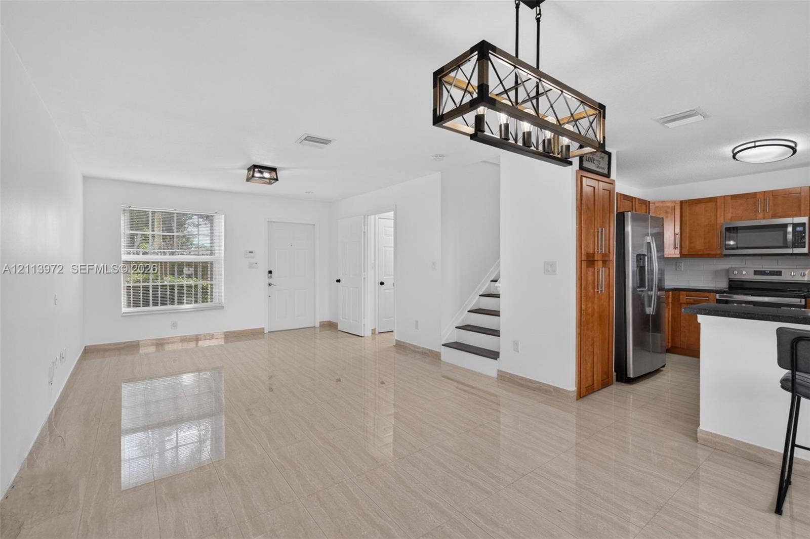 Large-format tile flooring, kitchen featuring wood cabinetry and stainless steel appliances, rectangular pendant lighting with metal frame, and staircase with dark-finish treads