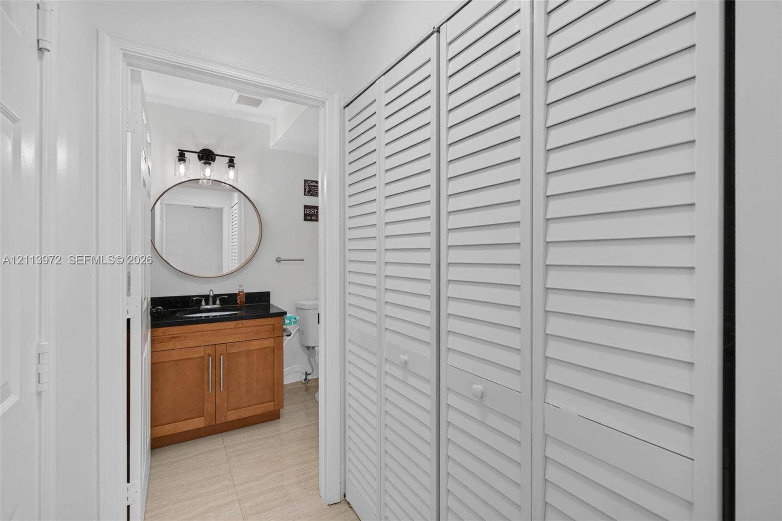 Built-in cabinetry featuring white louvered doors, wooden vanity with dark stone countertop, circular mirror, and multi-light vanity fixture