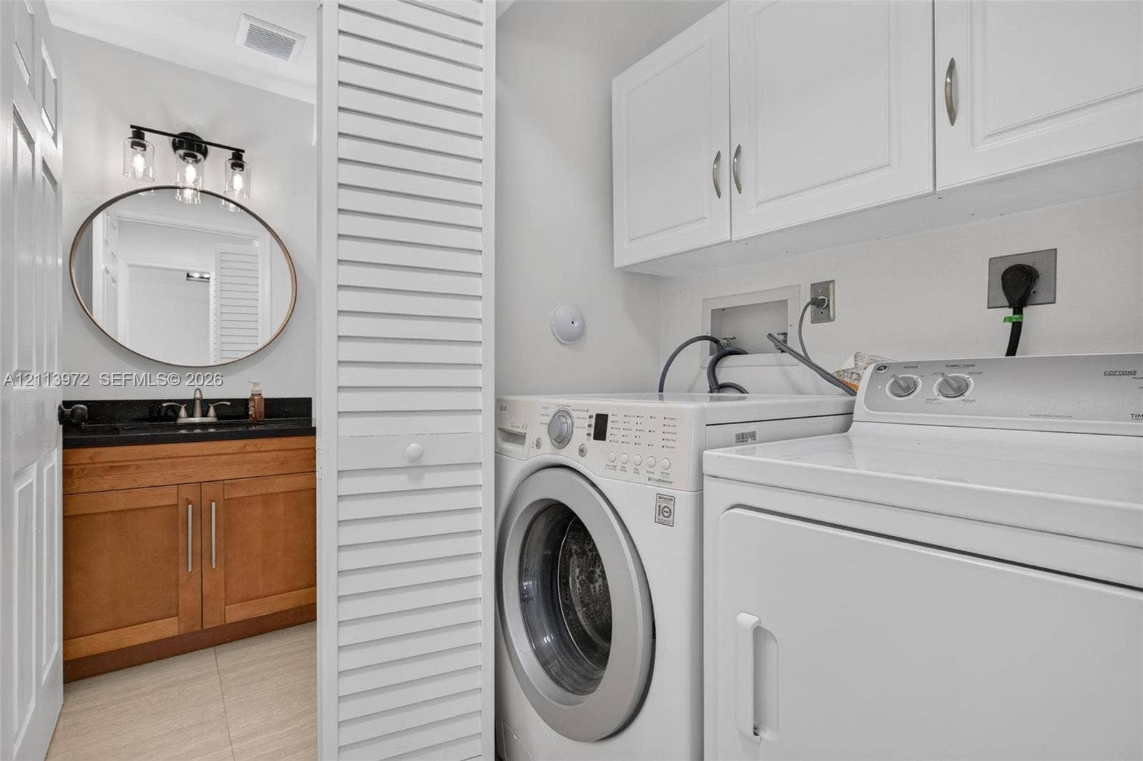 Laundry space featuring white upper cabinetry, louvered closet doors, and tile flooring, paired with a vanity area containing a wood-finish cabinet, dark countertop, round mirror, and three-light wall-mounted fixture