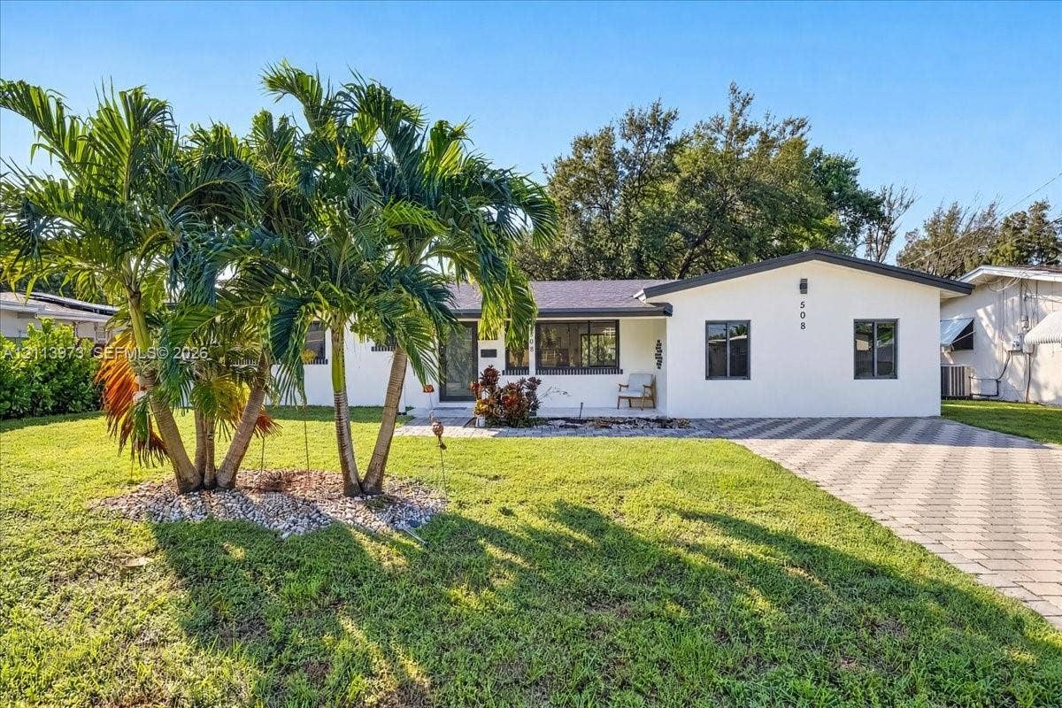 Single-story exterior featuring white stucco siding, a paved stone driveway, dark-trimmed window frames, and established palm tree landscaping