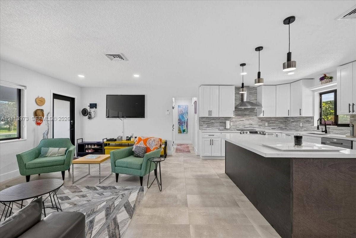 Kitchen featuring white cabinetry, an expansive island with a contrasting dark base, quartz countertops, a stone tile backsplash, and contemporary pendant lighting, set over large-format neutral tile flooring