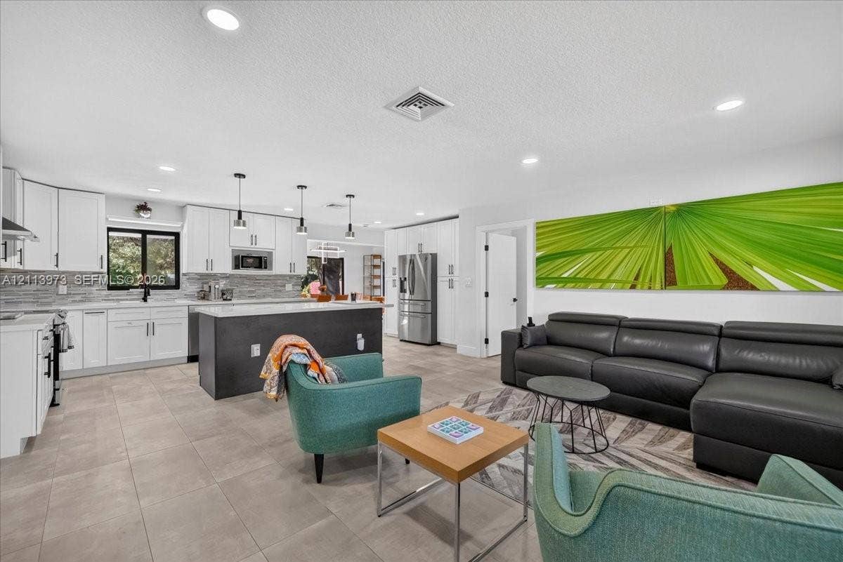 Open-concept kitchen featuring white shaker-style cabinetry, a contrasting dark central island, stone-tiled backsplash, stainless steel appliances, and large-format neutral floor tiling