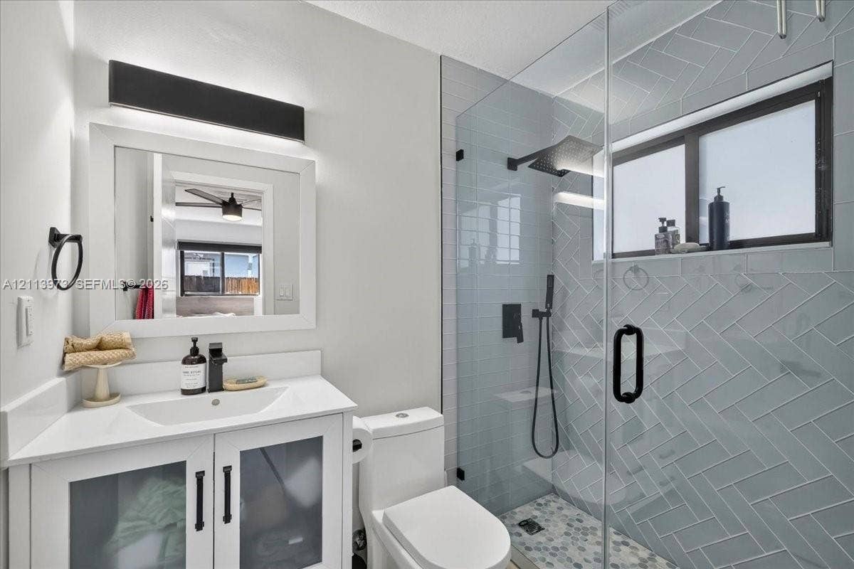 White vanity with a rectangular sink, herringbone-patterned wall tile, glass-enclosed shower with matte black fixtures, and custom light fixture