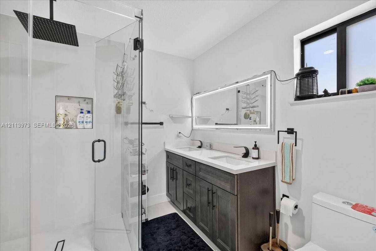 Double vanity featuring a light-toned countertop, dark-finished cabinetry, an illuminated mirror, a glass-enclosed shower with a rainfall showerhead, and a built-in recessed wall niche
