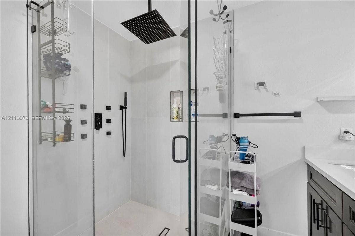 Walk-in shower featuring a frameless glass enclosure, oversized square rainfall showerhead, matte black hardware, integrated tiled niche, and light-toned wall tile