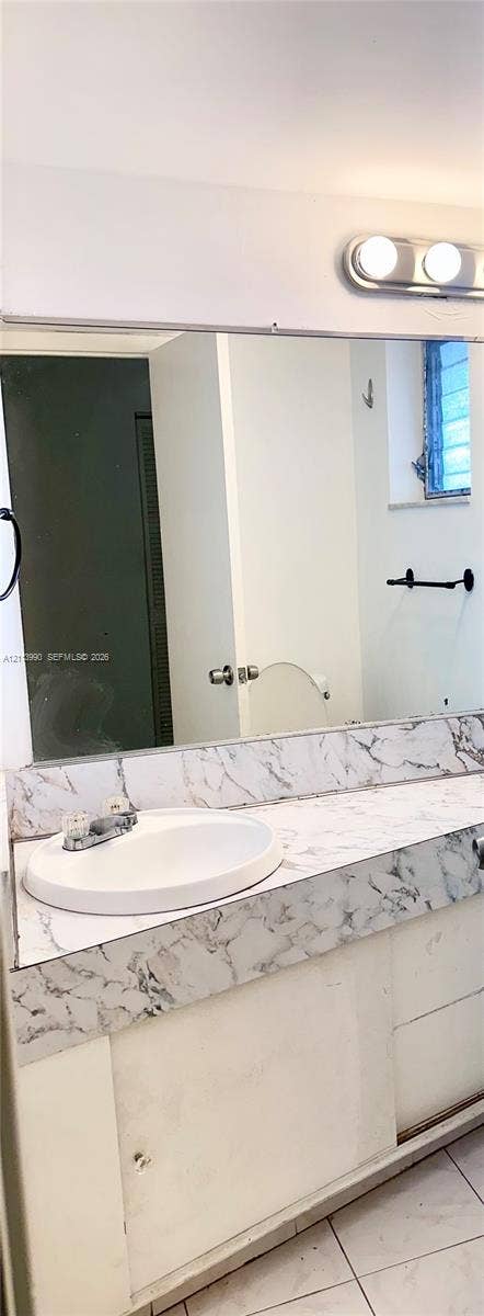 1501 NE 191st Street, #C312, Miami, FL 33179 Bathroom vanity featuring a stone-patterned countertop, an integrated sink with dual-handle faucet, full-width wall mirror, and dual-bulb vanity light fixture