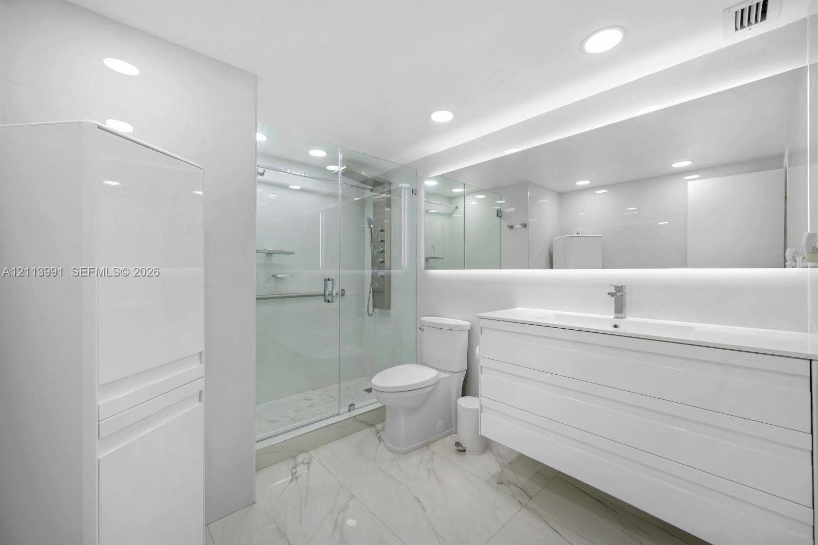 Modern bathroom featuring large-format tile flooring, a glass-enclosed shower with a multi-head body spray system, a floating vanity with a seamless integrated sink, and an expansive wall-to-wall mirror with integrated LED lighting