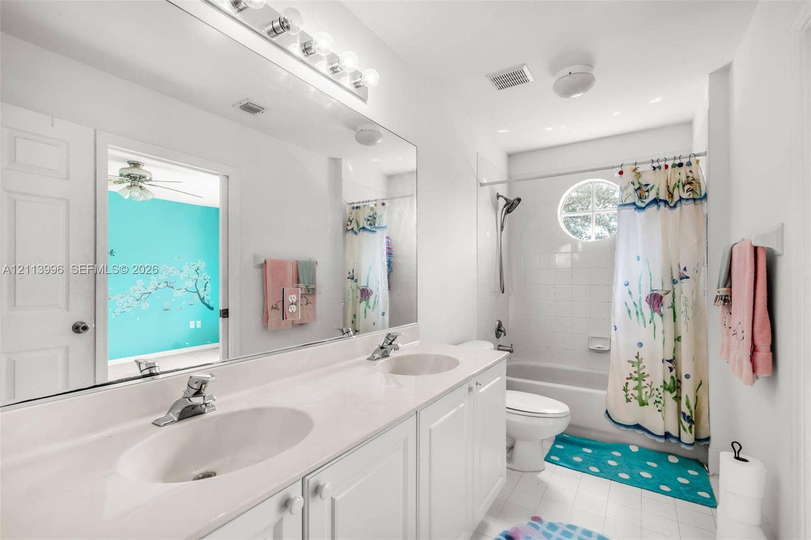 15503 SW 19th Street, Miramar, FL 33027 Double vanity with integrated white countertops and dual under-mount sinks, expansive wall-to-wall vanity mirror, overhead vanity lighting, circular accent window, and white tile tub surround
