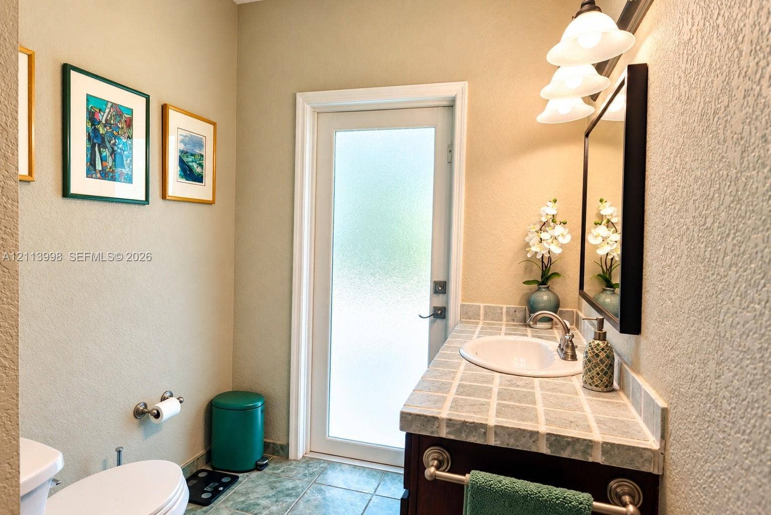 16455 SW 91st Avenue, Palmetto Bay, FL 33157 Vanity featuring a tiled countertop, undermount sink, and brushed metal faucet, complemented by a tri-light wall fixture, framed mirror, and exterior door with privacy glass