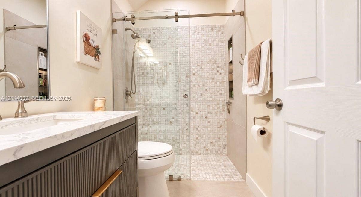 1495 NE 33rd Avenue, #107, Homestead, Homestead, FL 33033 Bathroom featuring a fluted wood-finish vanity with a stone-surface countertop, an integrated sink, a glass-enclosed walk-in shower with mosaic tile accents, and built-in wall shelving