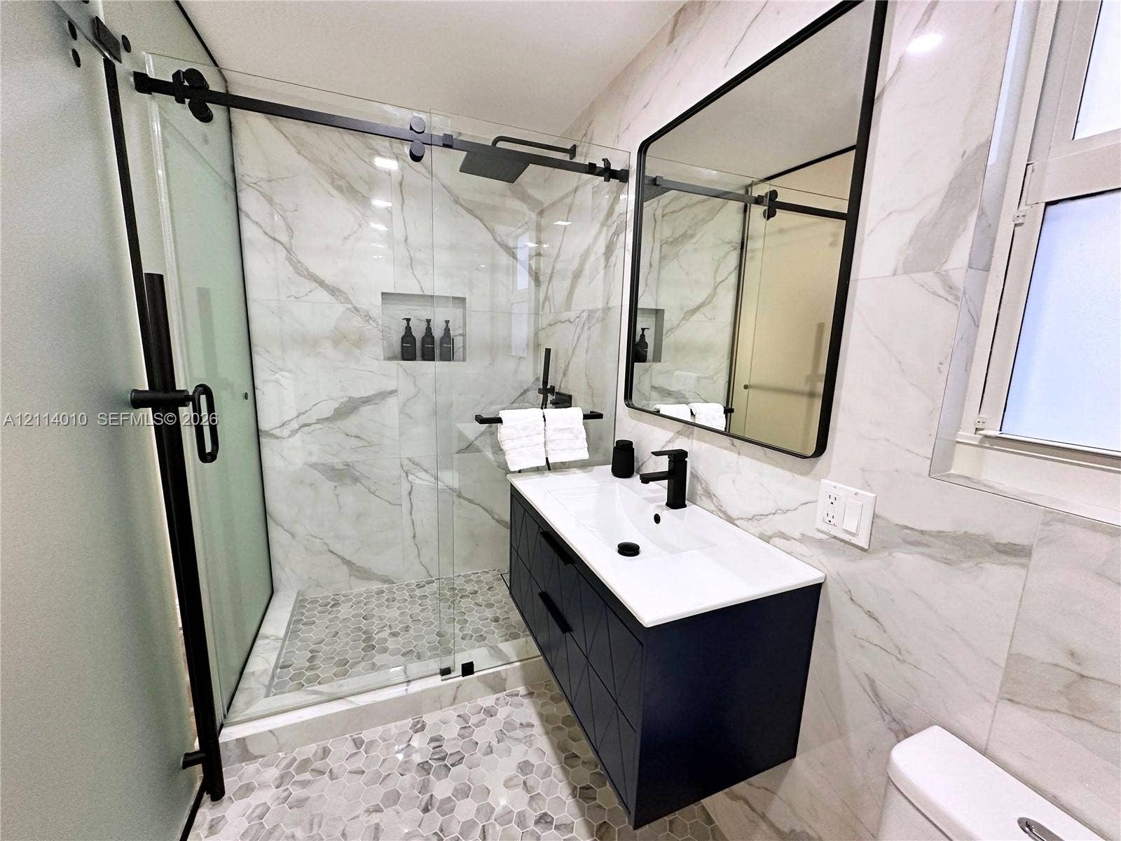 1161-1163 SW 30th Street, Fort Lauderdale, FL 33315 Walk-in shower featuring a sliding glass door, matte black hardware, integrated wall niche, and stone-tiled floor with a matching vanity and black-framed mirror