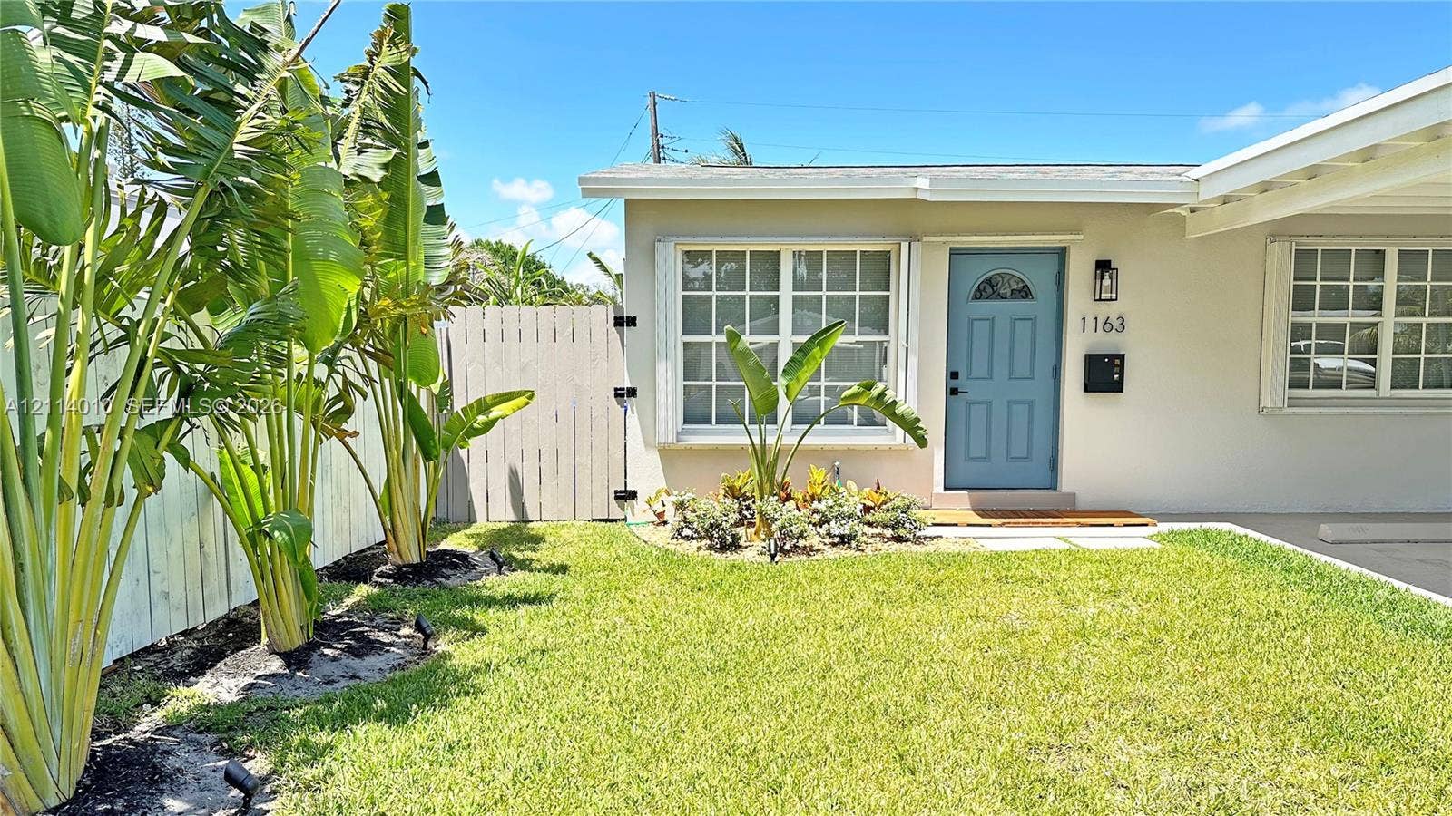 1161-1163 SW 30th Street, Fort Lauderdale, FL 33315 Exterior featuring light-toned stucco siding, a blue painted entry door, grid-pattern windows, an attached carport, and a fenced perimeter