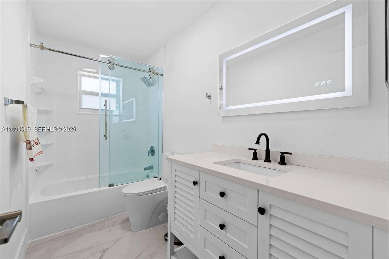 Bathroom featuring a white vanity with louvered cabinet doors, stone-composite countertop, illuminated wall mirror, and a glass-enclosed shower with chrome hardware