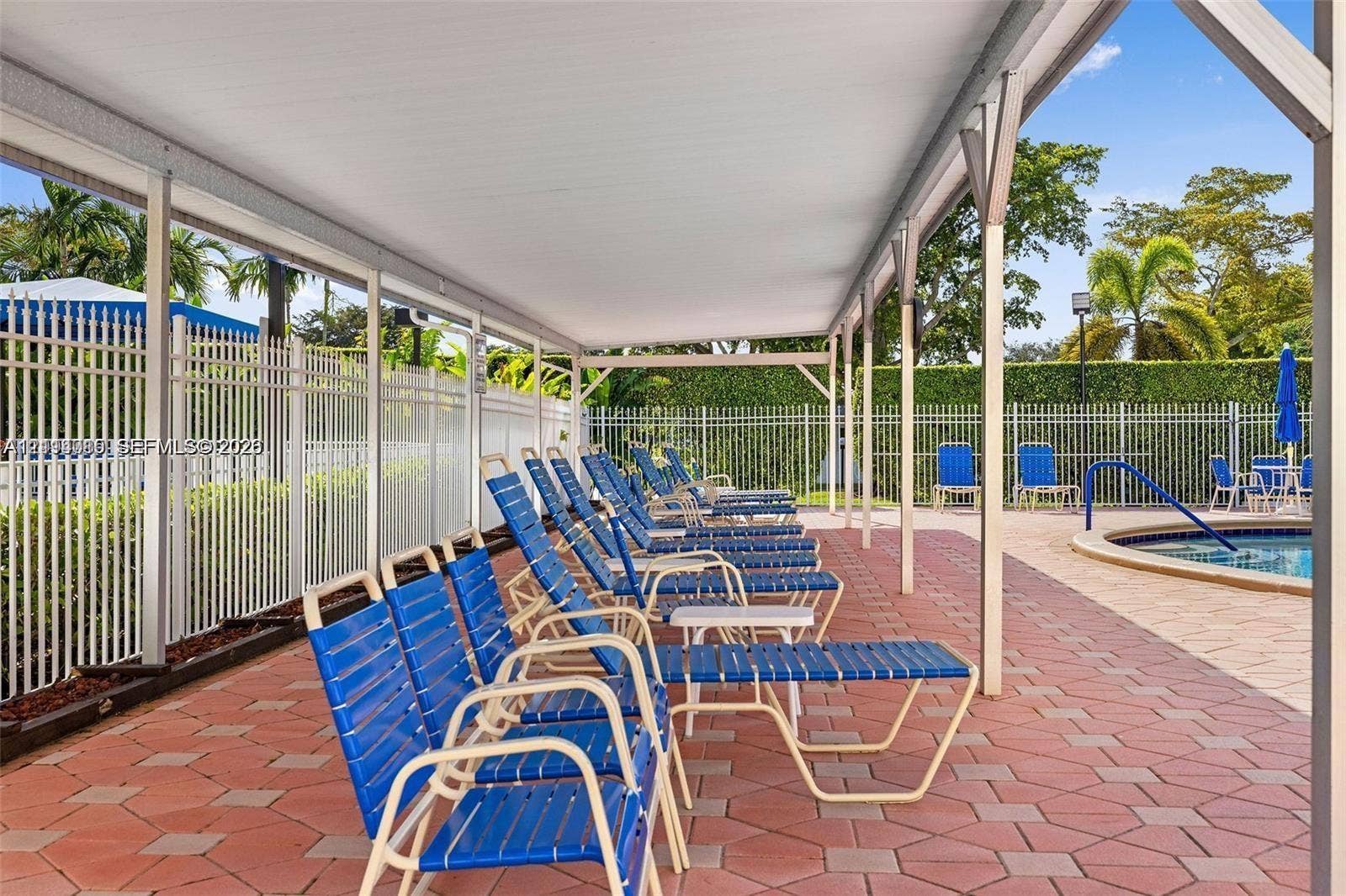 Covered outdoor patio with red paver flooring, white metal perimeter fencing, and swimming pool