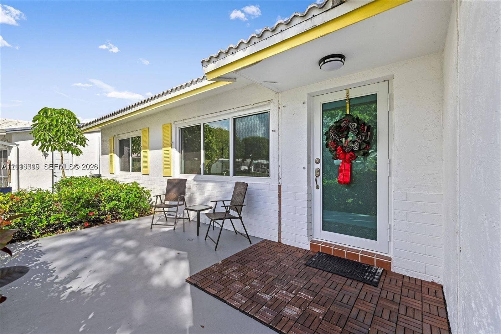 Exterior entryway featuring white painted brick masonry, yellow louvered shutters, decorative tile flooring, and a glass-paneled entry door