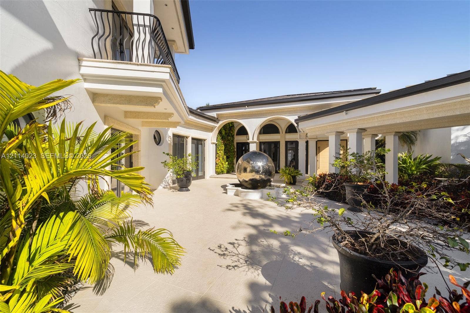 Courtyard featuring a central metallic sphere fountain, covered colonnade, arched architecture, exterior balcony with wrought-iron railing, and stone-finish paving