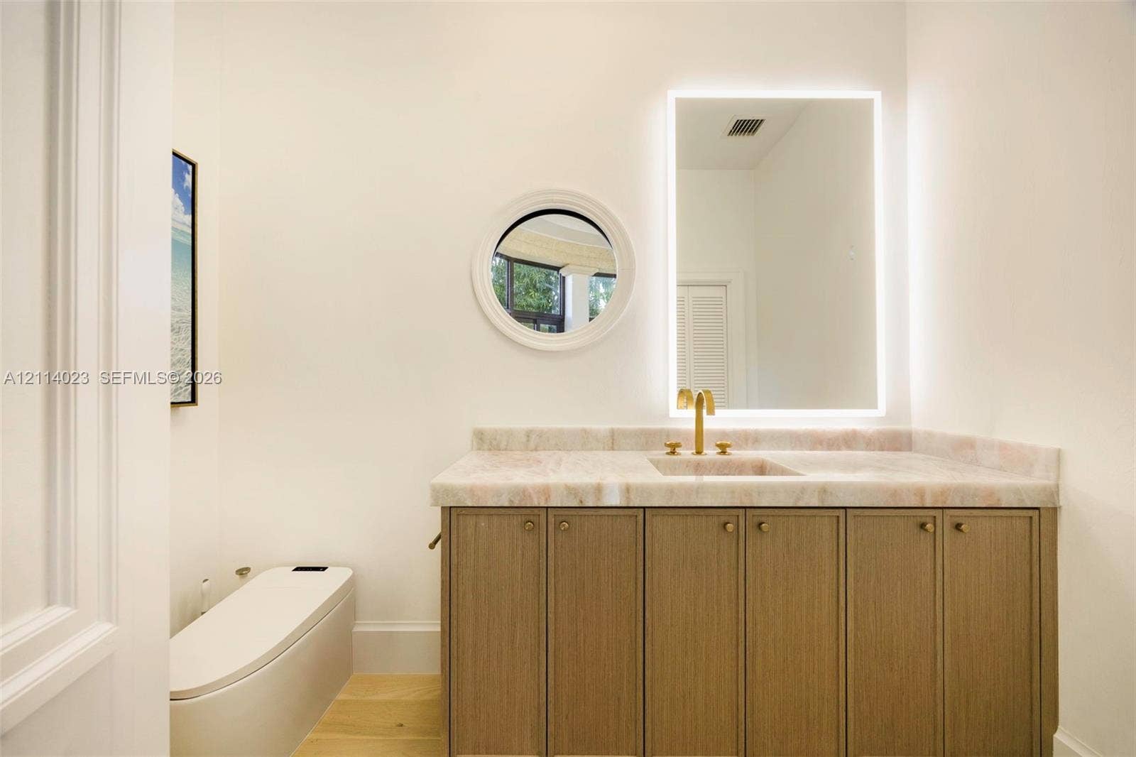 Custom wood-finish vanity with stone countertop, brass faucet, rectangular integrated sink, backlit LED mirror, and circular accent window